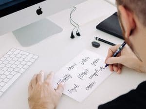 Person schreibt kreative Ideen auf Papier mit Computer im Hintergrund.